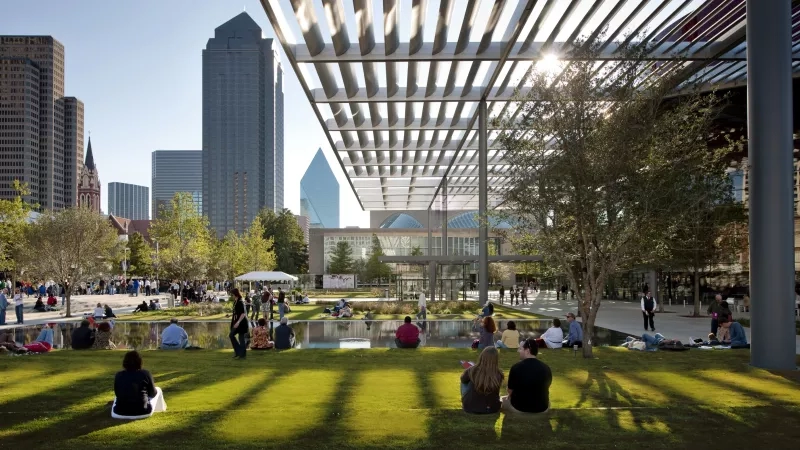 People enjoy the Dallas Arts District on a sunny day, with skyscrapers visible in the distance.