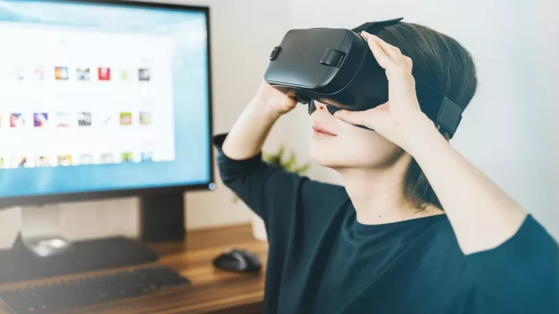 Woman using virtual reality headset with a monitor and keyboard in the background.