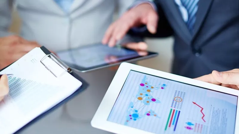 Three business people in a meeting with iPads and graphs on the screen.