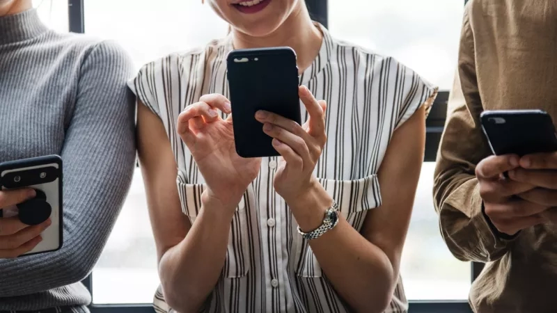 Three people in casual clothing holding cellphones in front of contemporary glass architecture.