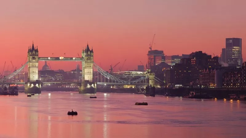 The London skyline with Tower Bridge, a body of water, and a pink sky at sunset.