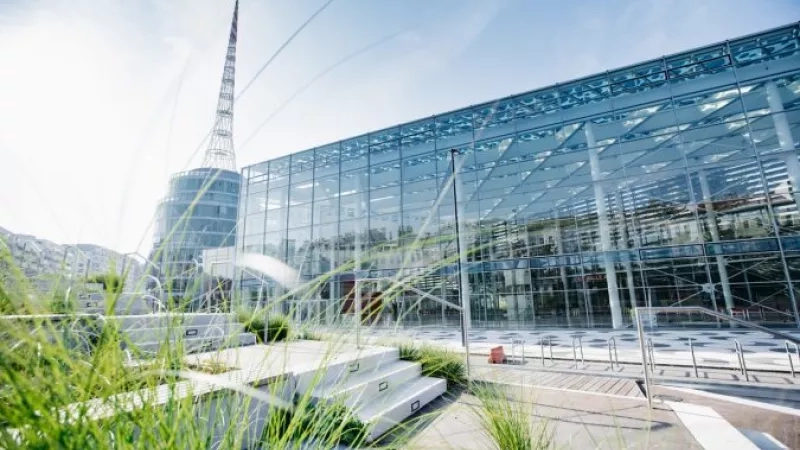 Modern glass building with steps, green plants, and a tower in the background under a clear sky.