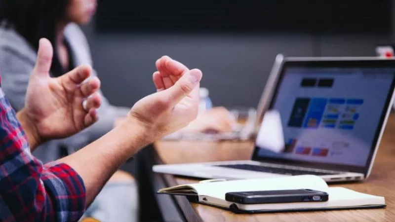 Person's hands gesturing in front of a laptop, with a notebook and phone on a wooden table.