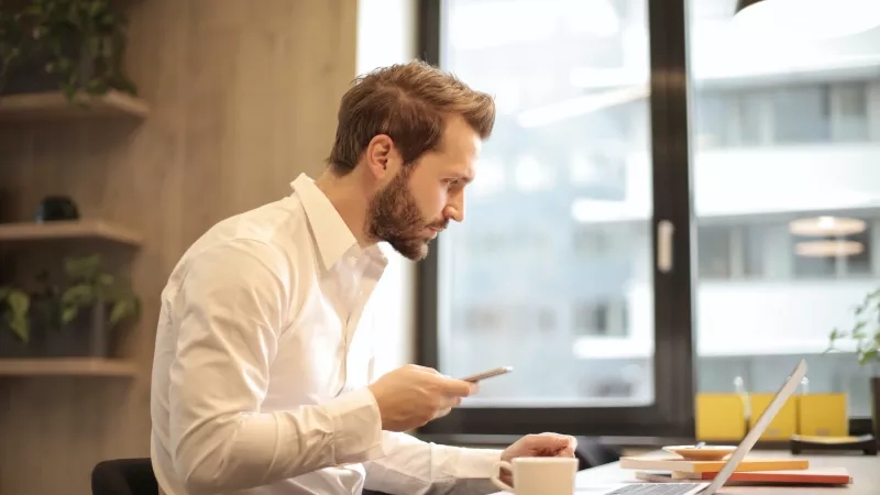 Man using cell phone and laptop while sitting in office with glass window in background.
