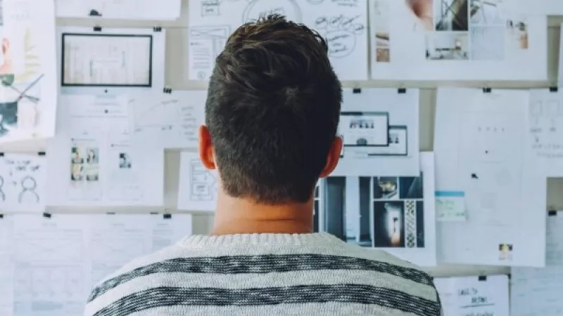 A man with a striped sweater looks at a noticeboard full of papers, architecture, and landscaping.