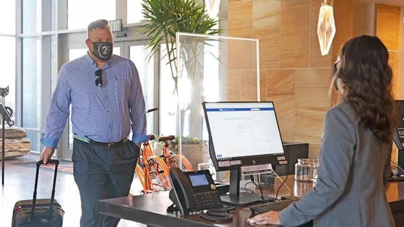 A man wearing a mask checks in at a desk with a computer monitor in the hotel lobby.