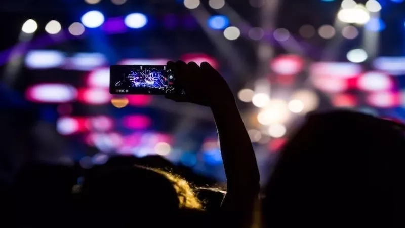 A person's silhouette is holding a smartphone at a live event with colorful lights and a stage.