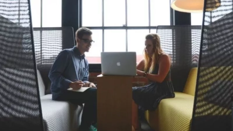 A man and a woman are sitting on a couch, working on a laptop on a table.