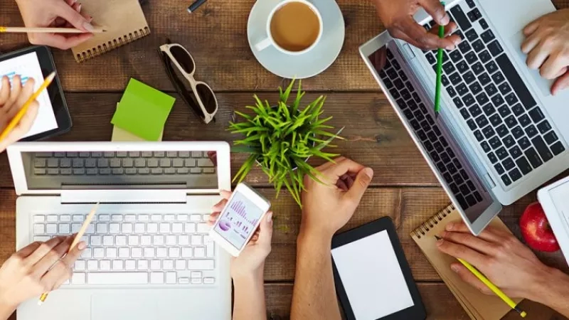 A group of people are gathered around a table with laptops, phones, and notepads, discussing event marketing tips.