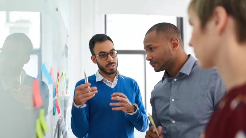 Group of people brainstorming ideas with sticky notes on a whiteboard in an office setting.