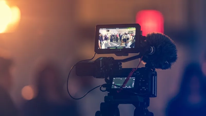 A tripod video camera with a microphone attached to it is set up in front of a crowd.