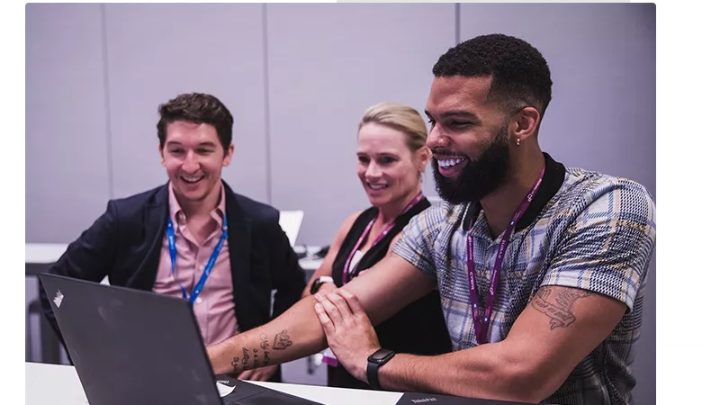 Three smiling people are working together on a laptop, showing teamwork and collaboration in a professional setting.