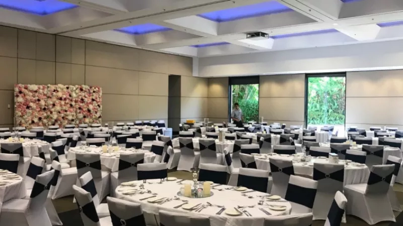 Banquet room at Novotel Twin Waters Resort with round tables, floral centerpieces, and a decorative wall.