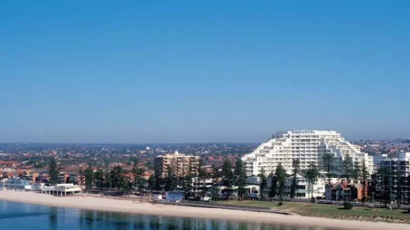 A view of Novotel hotel with beach on the left and buildings on the right with blue sky.