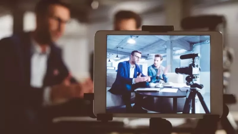 Two men sitting at a table on a tablet screen with a video camera in front of them.