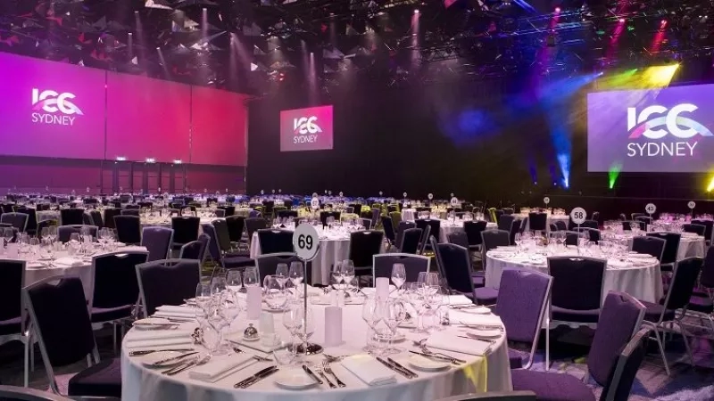 Banquet hall of ICC Sydney with white tables, chairs, glasses, and purple lights on the wall.