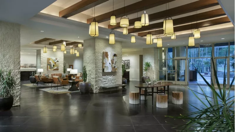 The lobby of a hotel with a table and chairs, plants, and hanging lights on the ceiling.