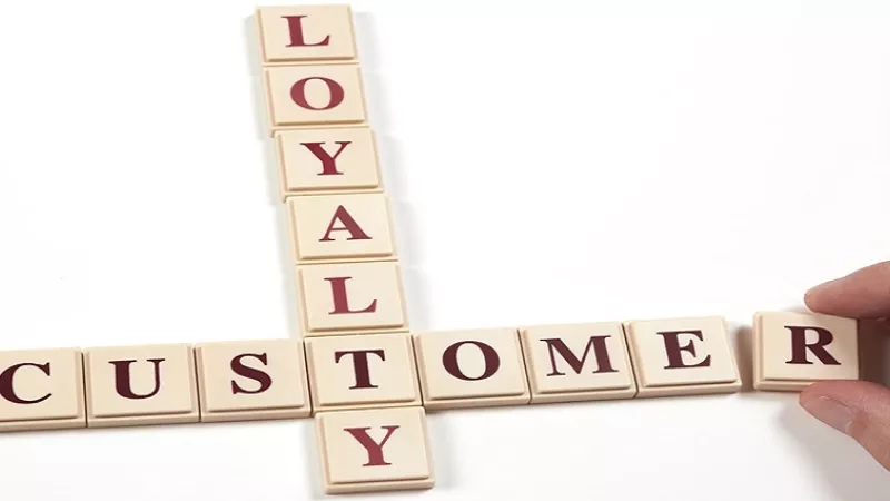 A hand holds a Scrabble tile to spell "customer loyalty" in beige tiles on a white background.