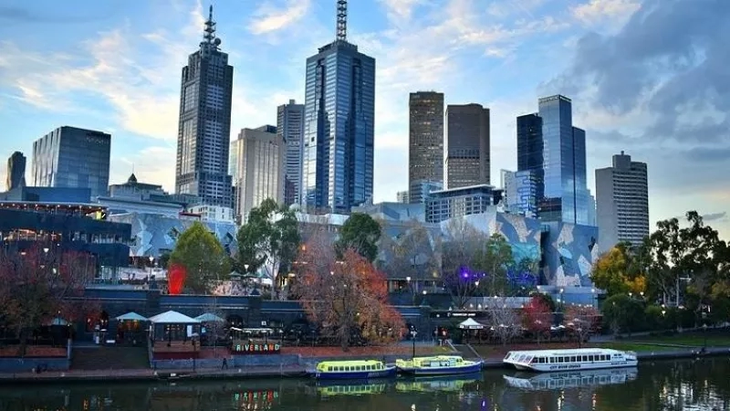 Melbourne city skyline with buildings, trees, and boats on a river under a cloudy sky.