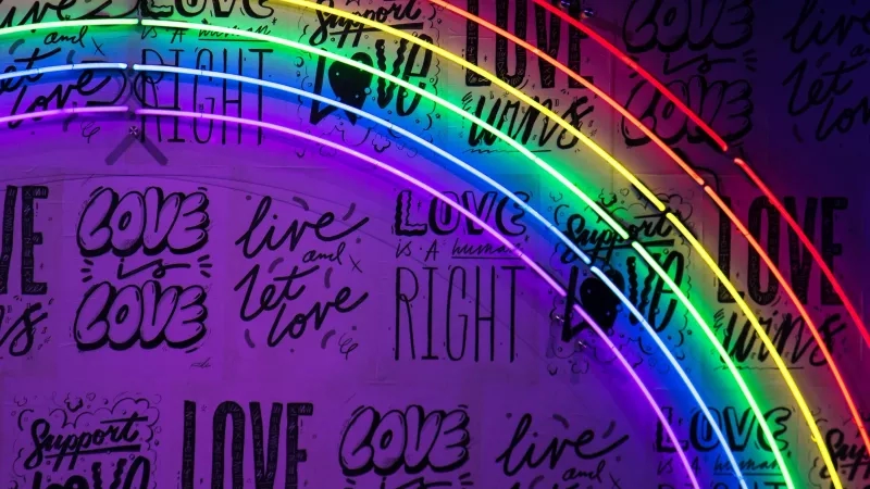 A rainbow-colored neon light is mounted on a purple wall that is covered in love messages.