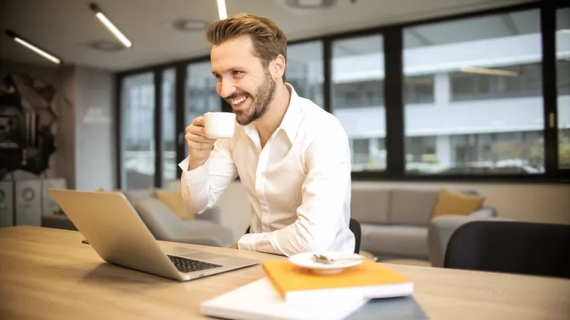Man smiling and drinking coffee while sitting at a table with a laptop in front of him.