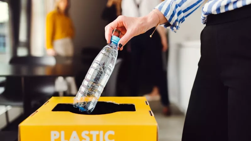 Woman putting a plastic bottle in a recycling bin with other people in the background.