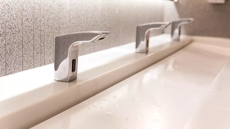 A close-up of a touchless faucet and sink in a restroom with a mirror in the background.