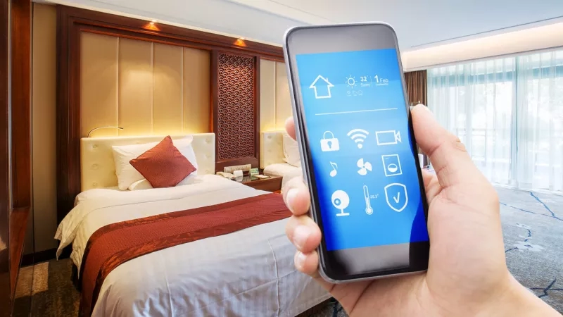 Hotel guest holding smartphone with smart home app icons on the screen in front of bed.