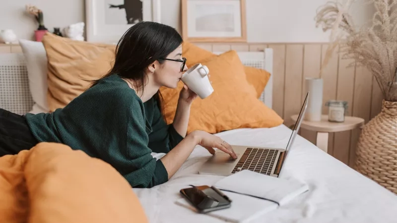 Woman in bed with a laptop drinking coffee during a virtual social event.