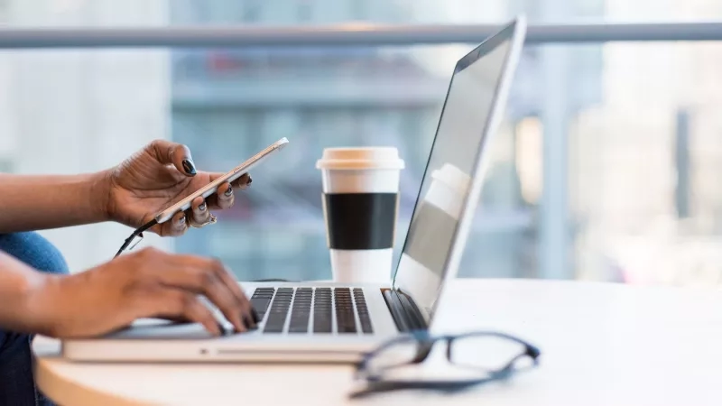 Person with phone and laptop on a table with a coffee cup and glasses.