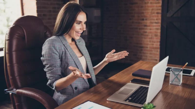 Woman in business suit sitting in an office chair at her desk with a laptop and paperwork.