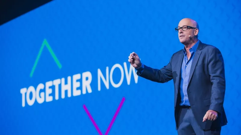 A speaker is giving a presentation on stage in front of a large blue screen that says "Together Now."