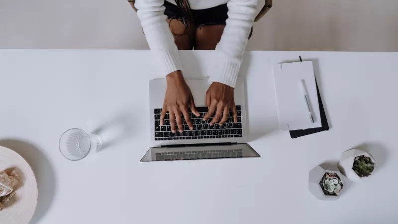 A woman typing on a laptop surrounded by plants and a notebook on a desk.
