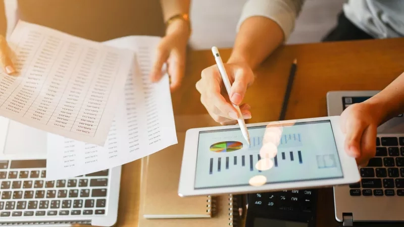 A person is reviewing financial information on a tablet while holding a pen and paper.