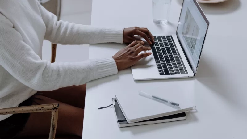 Person typing on laptop in home office with notepad and pen on desk in front of them.