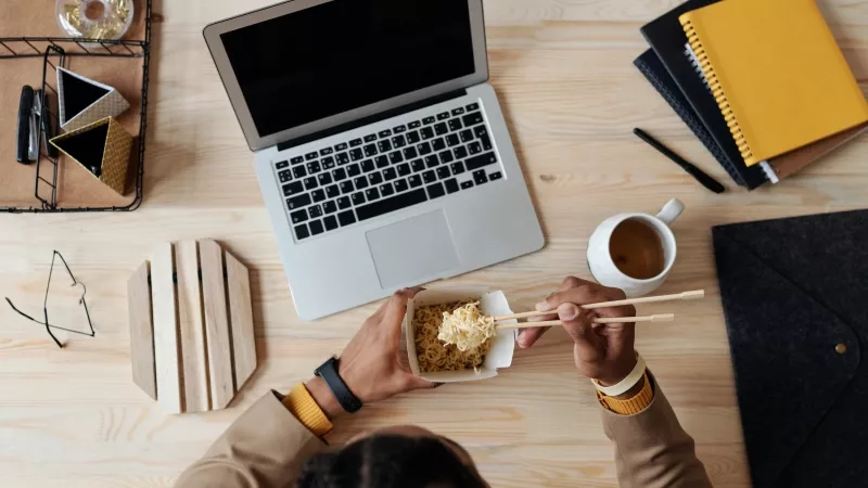 A person eats food using chopsticks in front of a laptop on a desk.