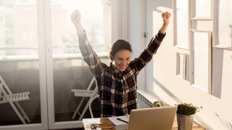 A woman in a plaid shirt is raising her hands while sitting at a desk with a laptop.