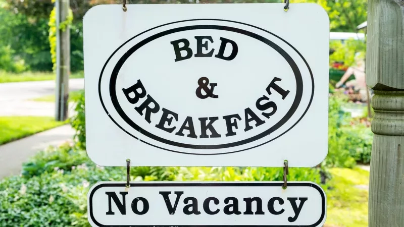 "Bed and Breakfast" sign hanging on a hook, with "No Vacancy" sign below, surrounded by greenery.