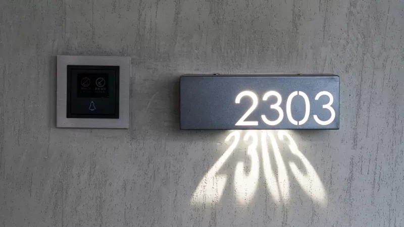 A wall-mounted room number with a lighted number 2303 and a light reflecting on the wall.