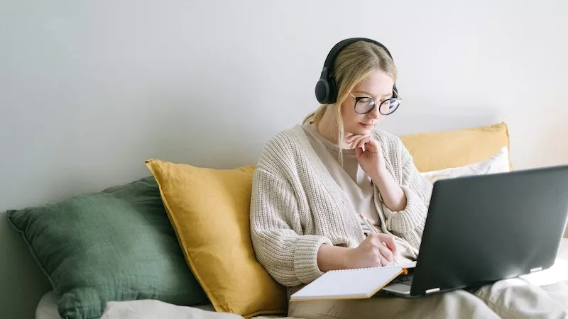 Woman with headphones and a laptop on a bed, taking notes in a notebook.