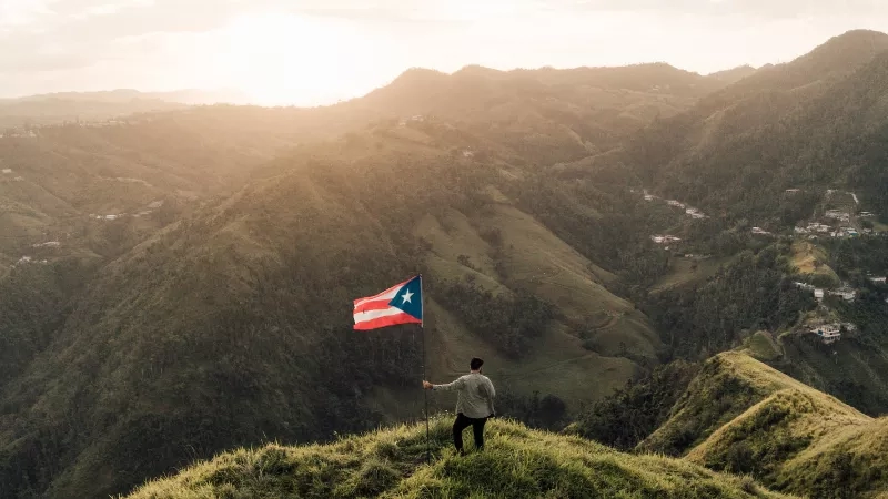 A man holding a Puerto Rico flag on a mountain overlooking a town in the valley.
