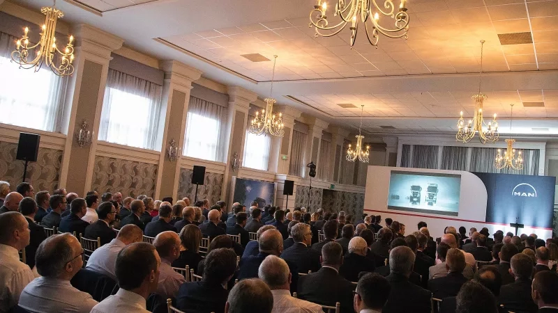 A large group of men gathered in a room with chandeliers, curtains, and a large screen.