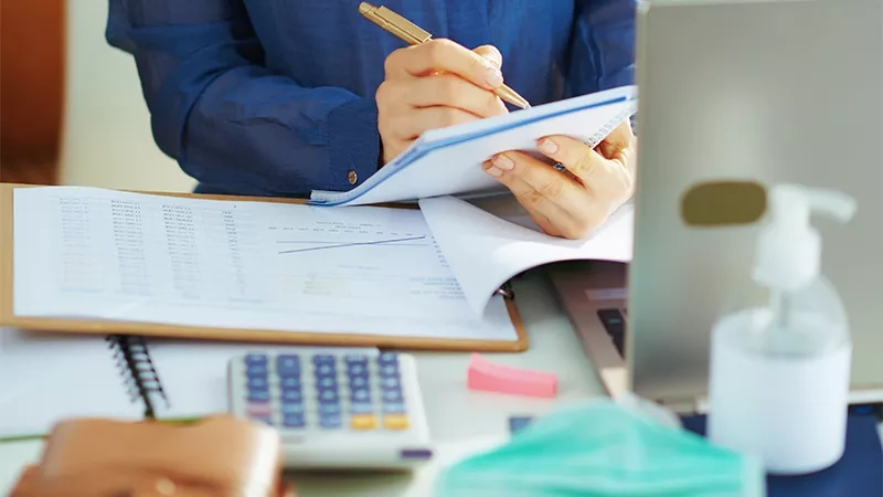 A woman is writing on a notepad with a pen while holding papers and a calculator on the table.