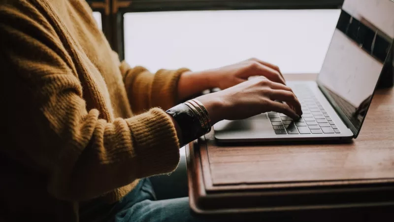 Person typing on laptop with one hand on keyboard and other on touchpad with natural lighting.