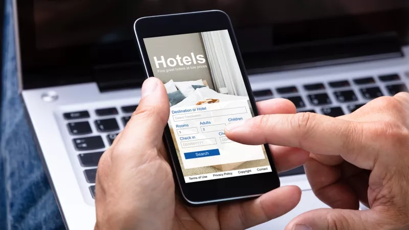 A person is using a mobile app to search for hotels on a laptop in a cozy room.