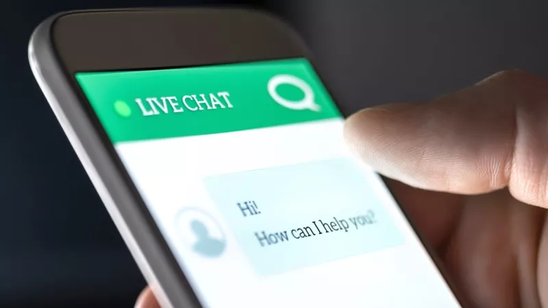 A person holding a mobile phone showing a live chat on the screen saying "How can I help you?"