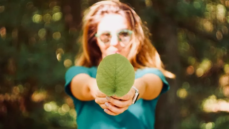 Woman in blue shirt holding up a leaf in front of her face in a forest.