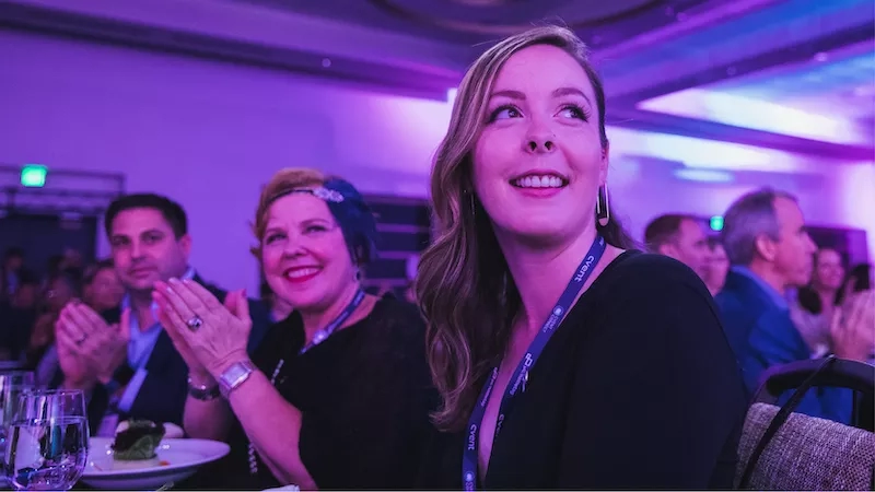 Three people are sitting in an event, with a woman in front wearing a lanyard with the text "CARE".