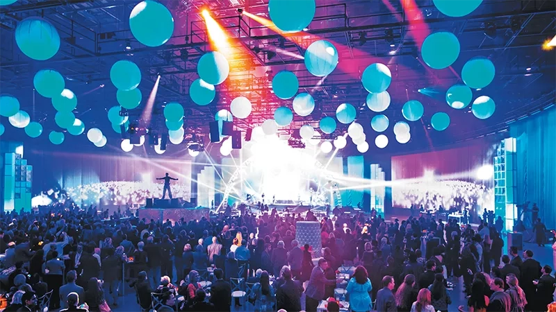 Crowded stage with people, balloons, and spotlights at a party in a large hall.