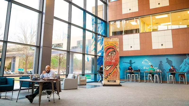 A man sitting at a table in a large, modern lobby with art, windows, and people.